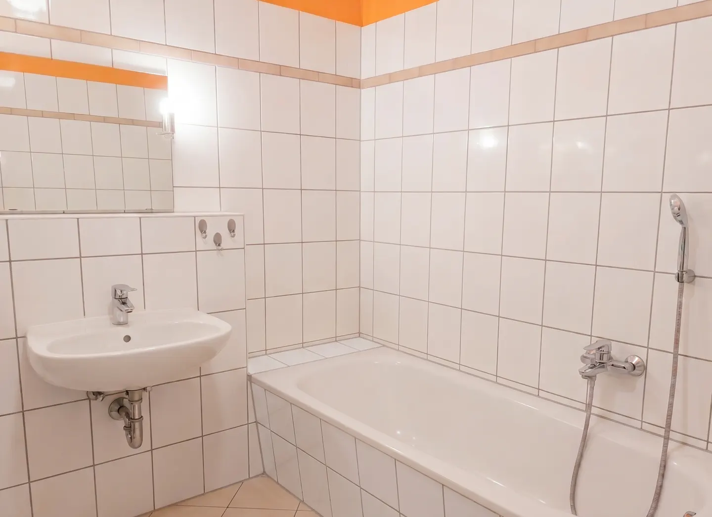 Bright bathroom with white square tiles, orange trim, a white sink, mirror, and a white bathtub with a shower head.