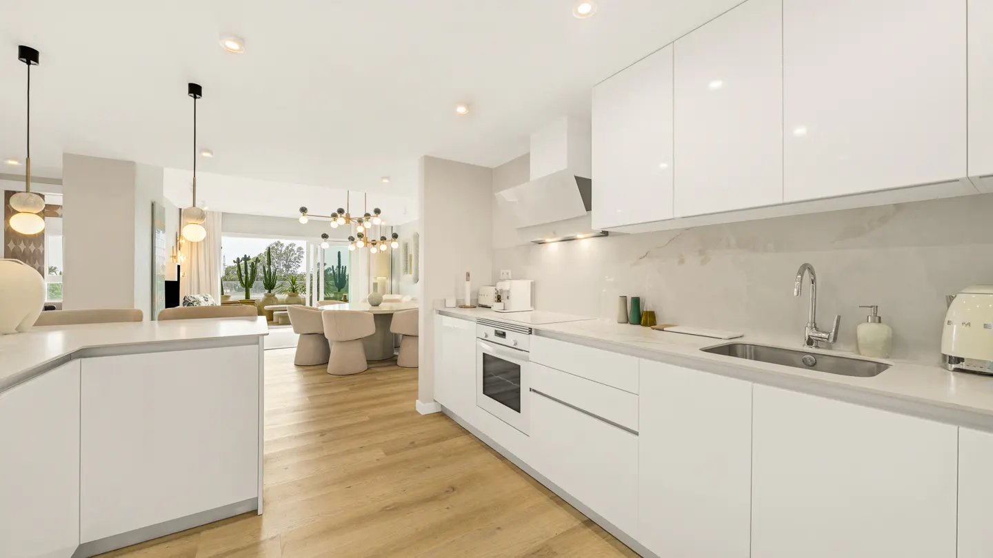 Bright, modern kitchen with white cabinets, marble countertops, and wood floors. Dining area visible in the background.