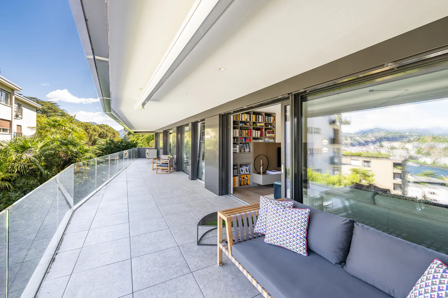 Wide balcony with gray sofa, patterned pillows, and glass railing overlooking trees and city. Sliding glass doors lead to a room with a bookshelf.