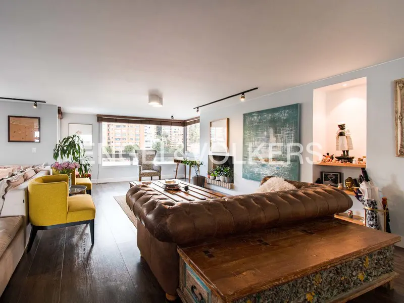 A living room with a brown leather sofa, a yellow chair, and a wooden coffee table. There is a window with a view of the city in the background.