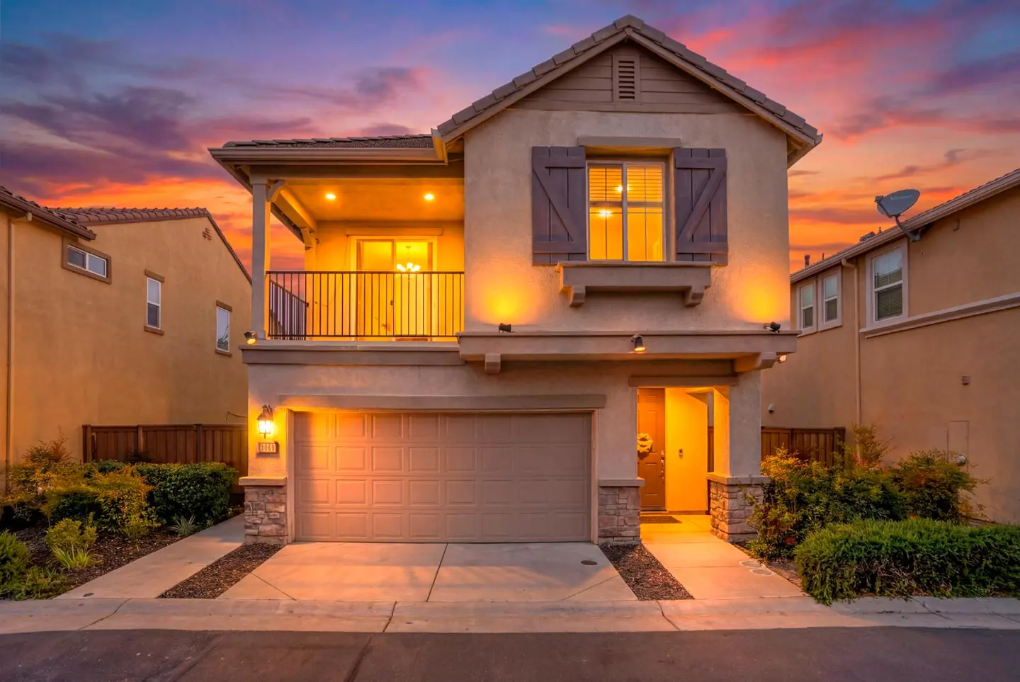 Two-story beige house with a balcony, brown shutters, and a garage at dusk with a colorful orange and purple sky.