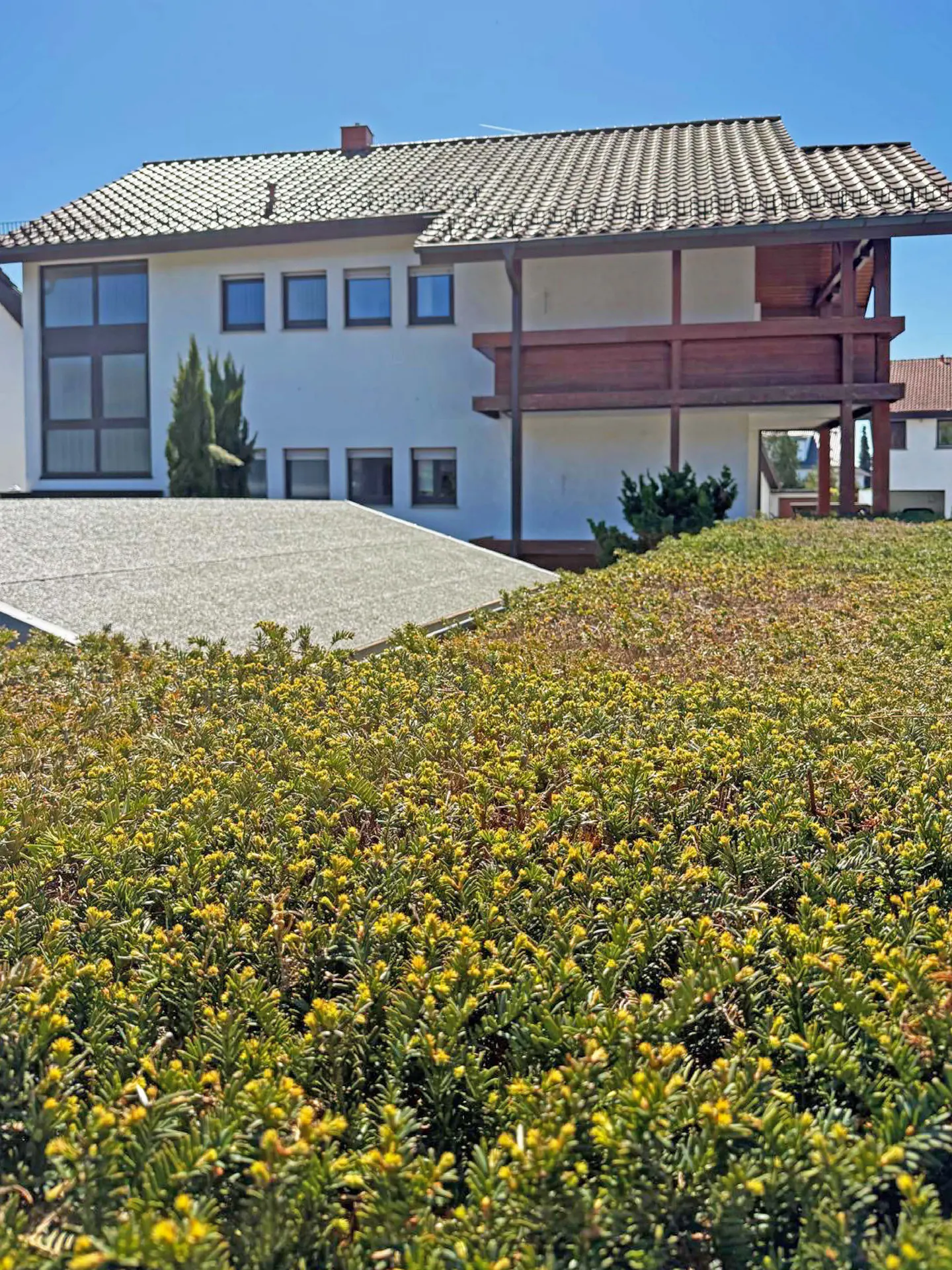 Two-story white house with a brown balcony and a gray tiled roof, viewed from behind a green and yellow bush.