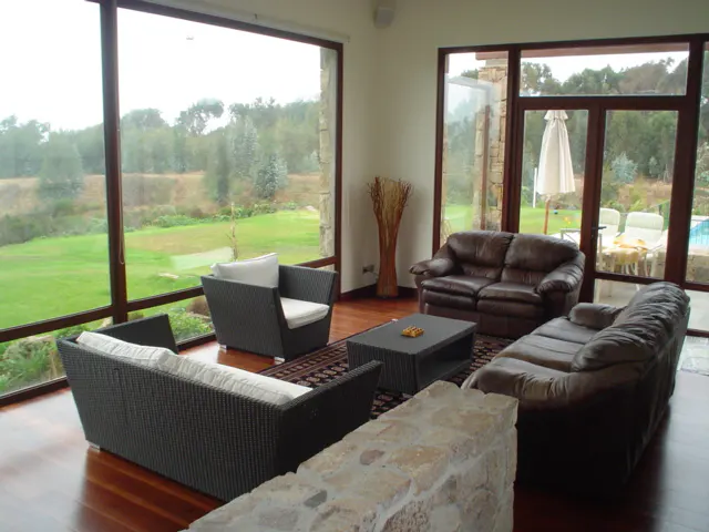 Living room with brown leather sofas, wicker chairs, and a black coffee table on a patterned rug. Large windows overlook a green lawn and trees.