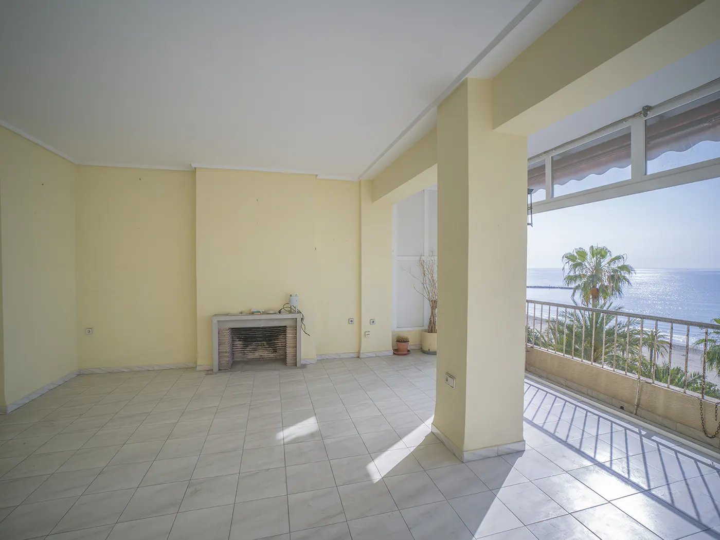 Bright, empty room with yellow walls, tile floor, and fireplace. Balcony with ocean view and palm trees visible through the open doorway.