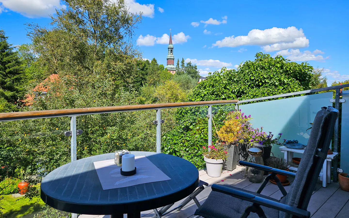 Balcony view with a round table, chair, and potted flowers. A church steeple rises above the trees in the background under a blue sky.