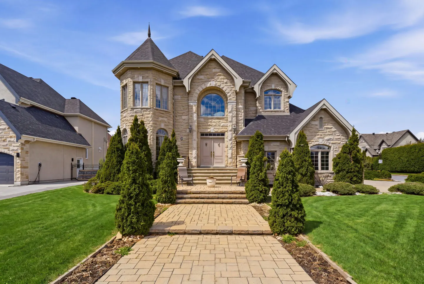 A large, two-story stone house with a turret, a brick walkway, and green lawn under a blue sky.