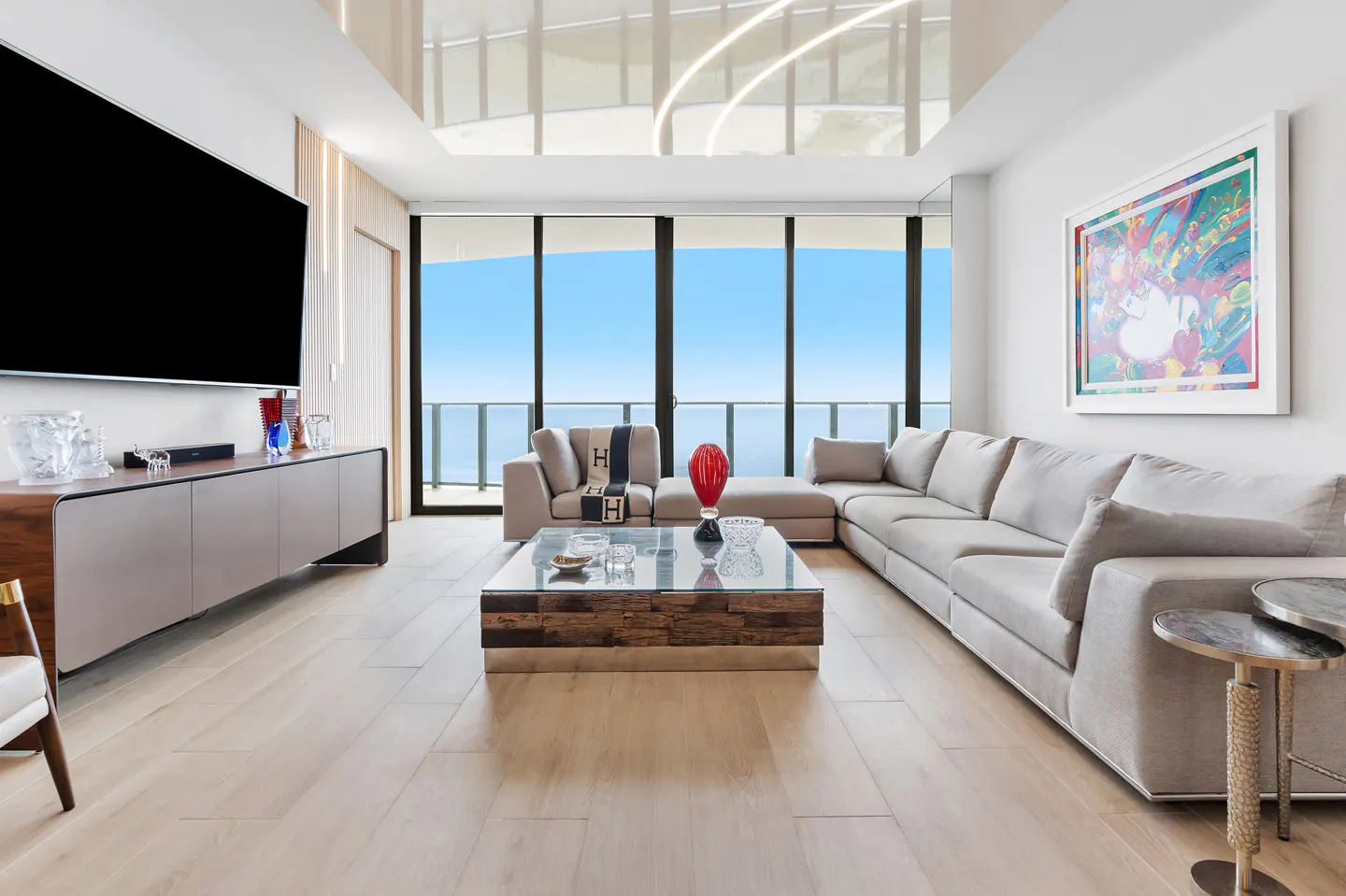 Bright living room with a large gray sectional sofa, glass coffee table, and ocean view through floor-to-ceiling windows. A large TV is mounted on the wall.