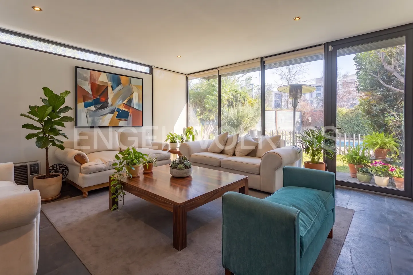 Bright living room with cream sofas, a teal armchair, a wooden coffee table, and large windows overlooking a garden.