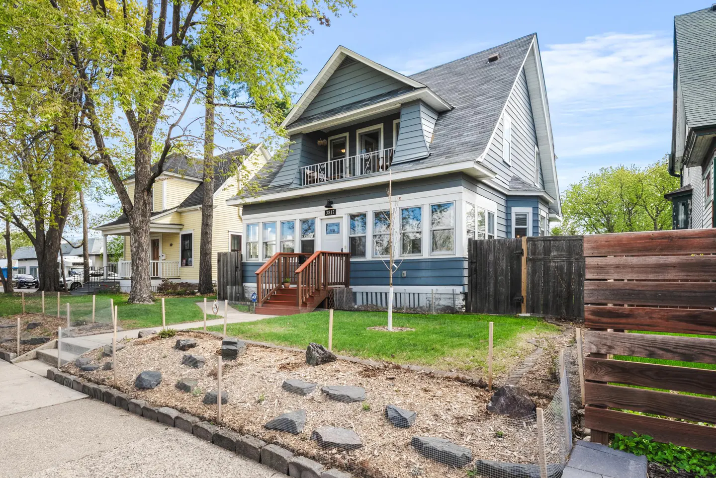 Two-story blue house with a gray roof, white trim, and a small balcony above the porch. A yellow house is next door.