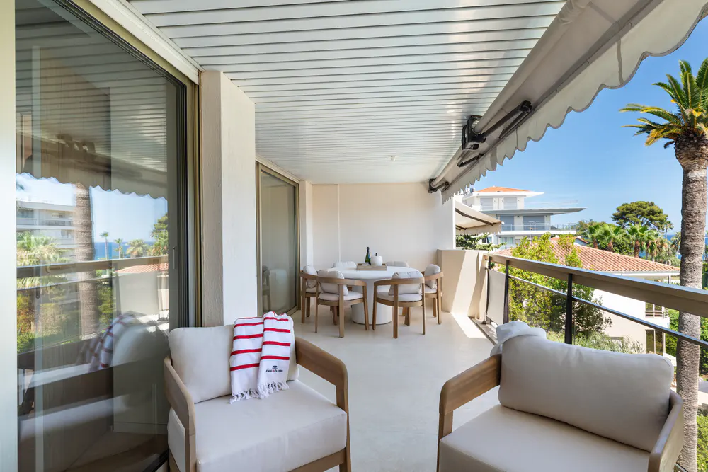 Covered balcony with white armchairs, a dining set, and a view of palm trees and nearby buildings under a clear blue sky.
