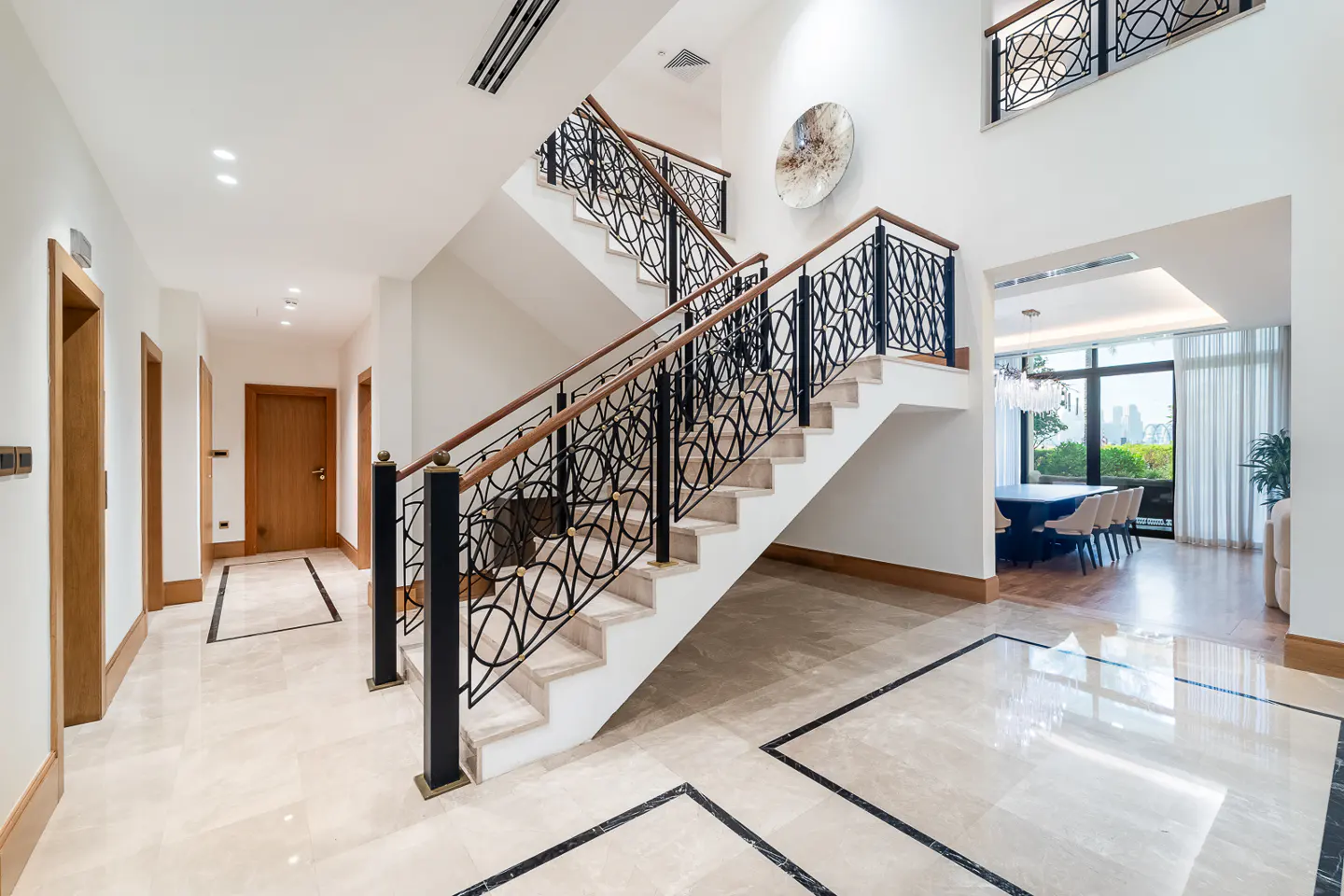 Bright foyer with marble floors, white walls, and a staircase with black wrought iron railings and wood handrails. Dining room visible in background.