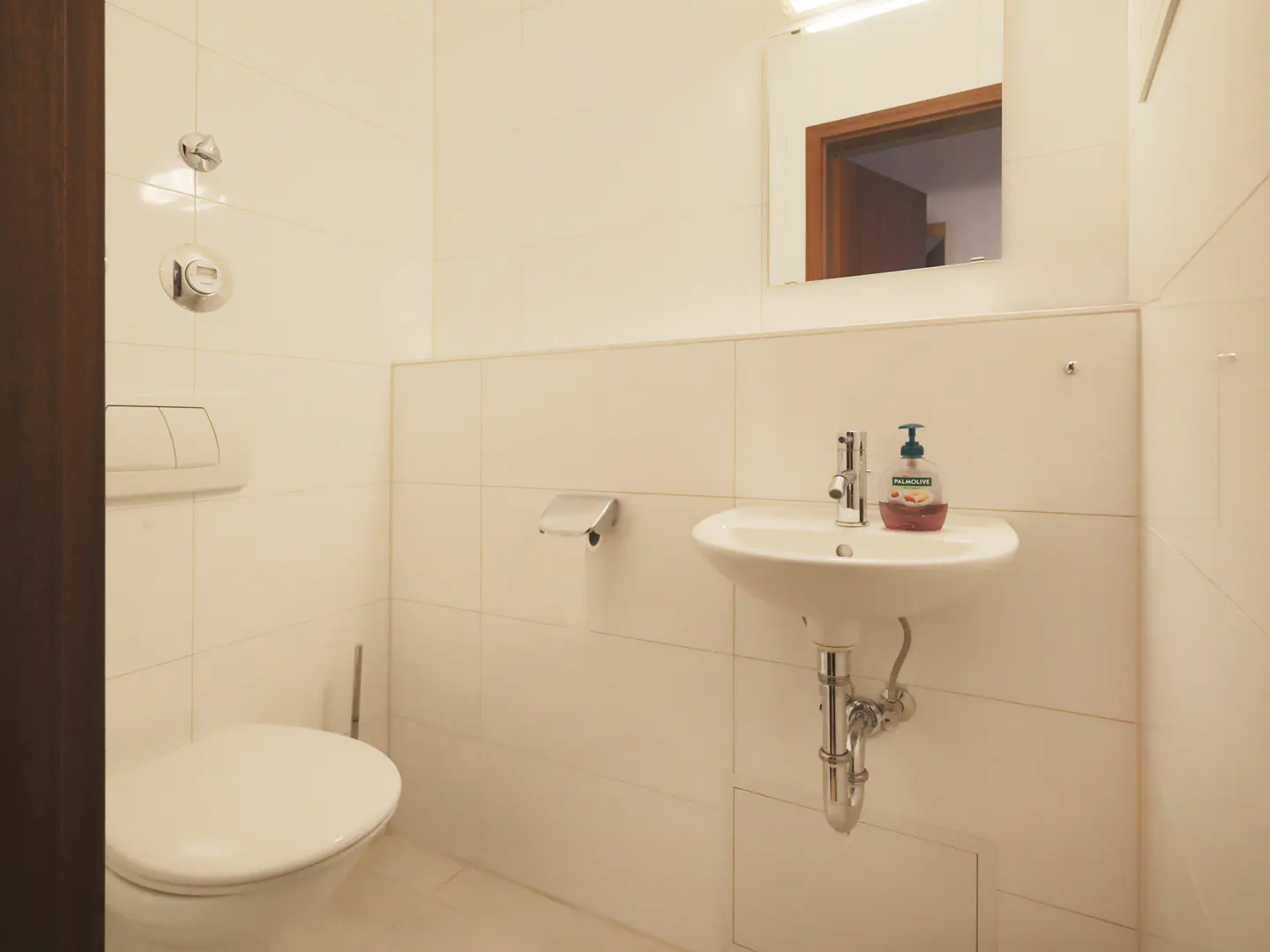 A small, white-tiled bathroom with a toilet, sink, mirror, and brown door frame.