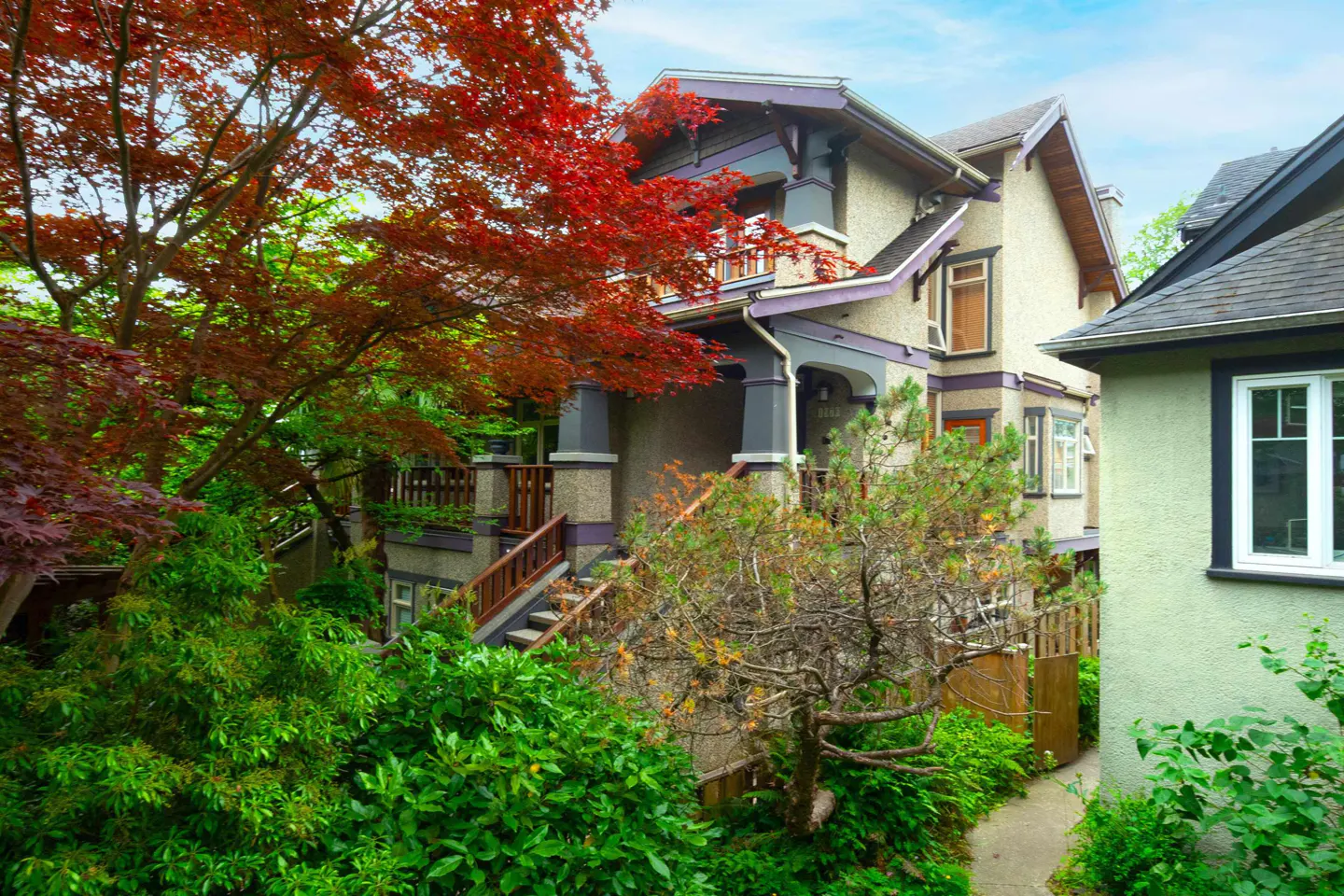 A multi-story house with beige stucco, dark trim, and a front staircase, surrounded by lush green trees and red foliage.