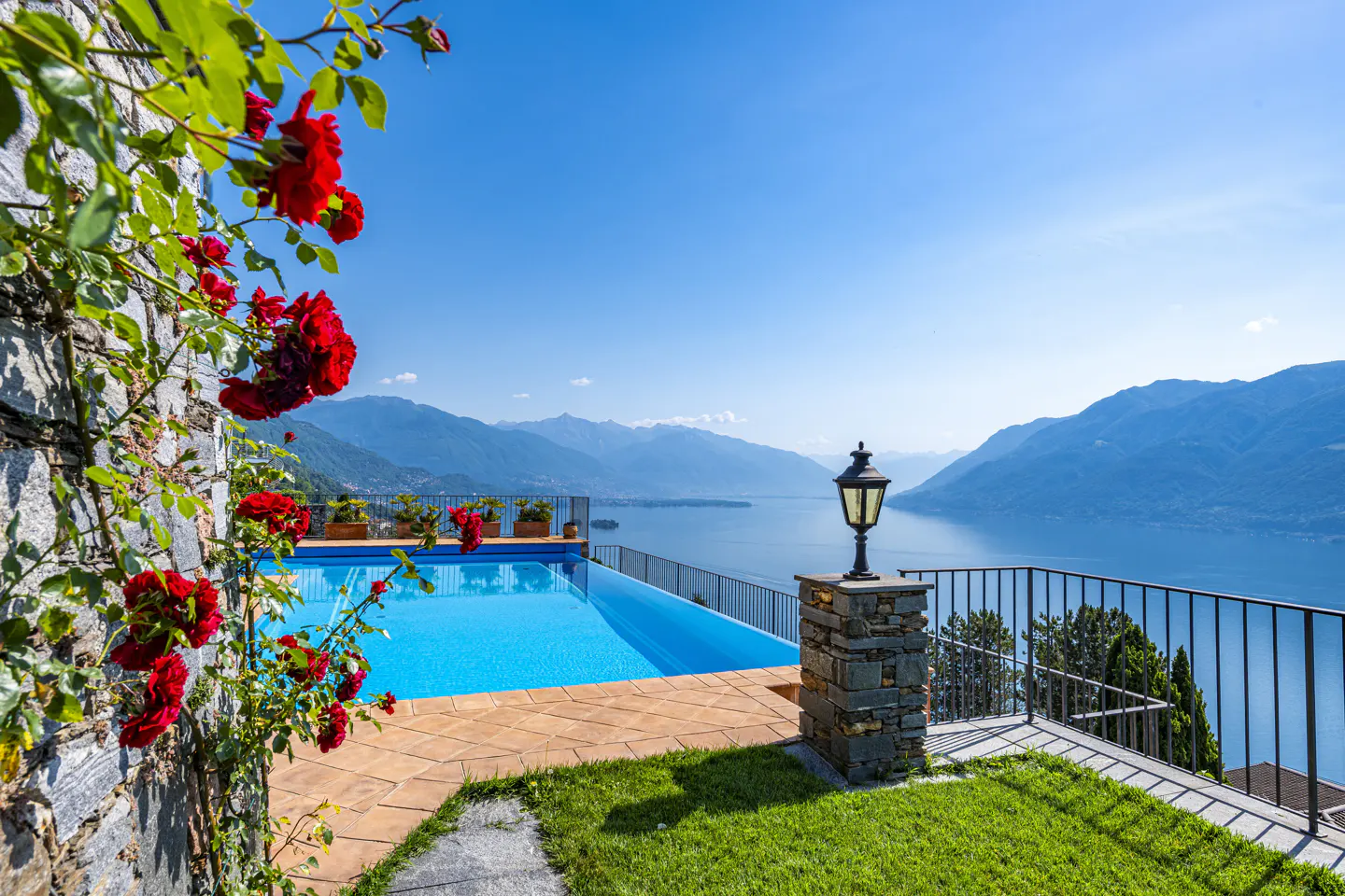 Infinity pool overlooking a lake and mountains, framed by a stone wall with vibrant red roses and a classic lamp post.
