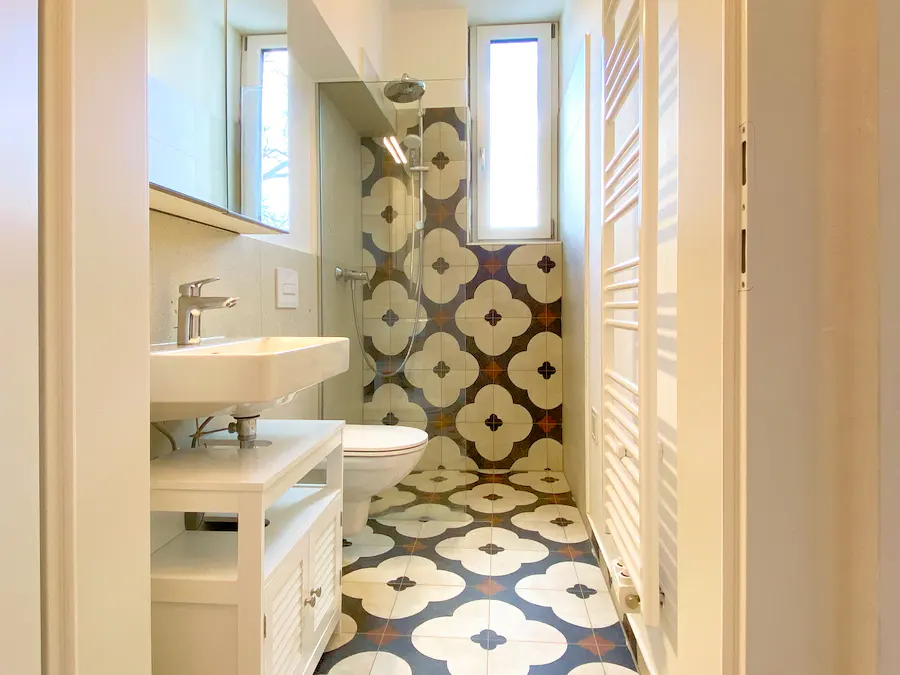 Bathroom with white sink, toilet, and shower. The floor and shower walls have patterned tiles. A white towel rack is on the right.