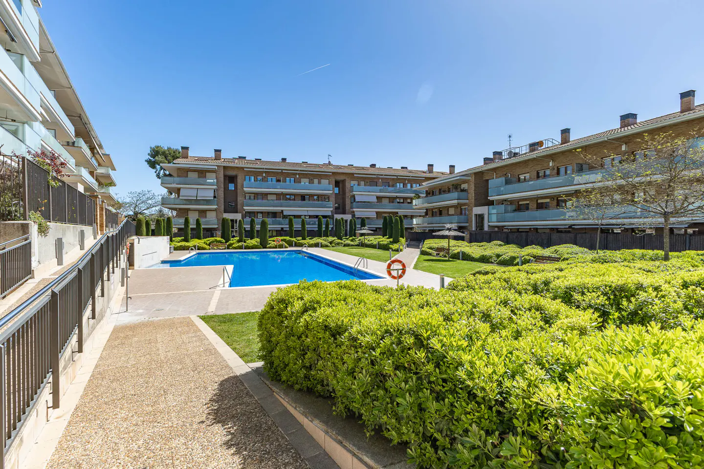 Apartment complex with a blue swimming pool, green bushes, and a clear blue sky. Balconies are visible on the apartments.