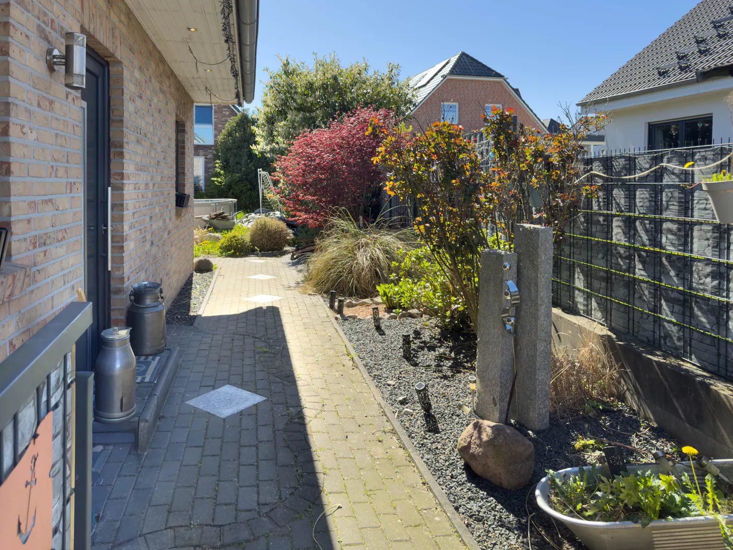 Brick house exterior with a stone walkway, garden, and fence on a sunny day. Two metal milk jugs sit near the front door.