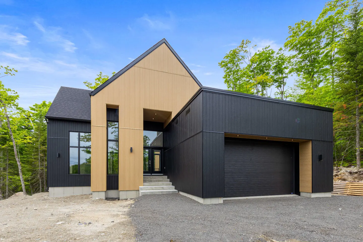Modern home with black and tan siding, a black roof, and a black garage door. The house is surrounded by trees and a blue sky.