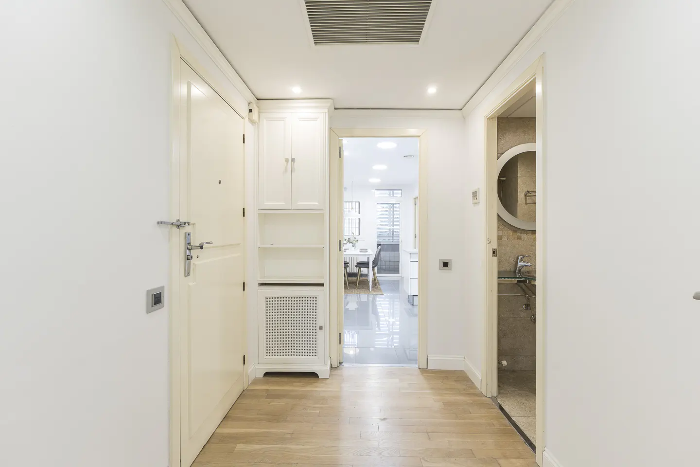 Hallway with light wood floors, white walls, and doors. A built-in cabinet is on the left. A bathroom is visible through an open doorway.
