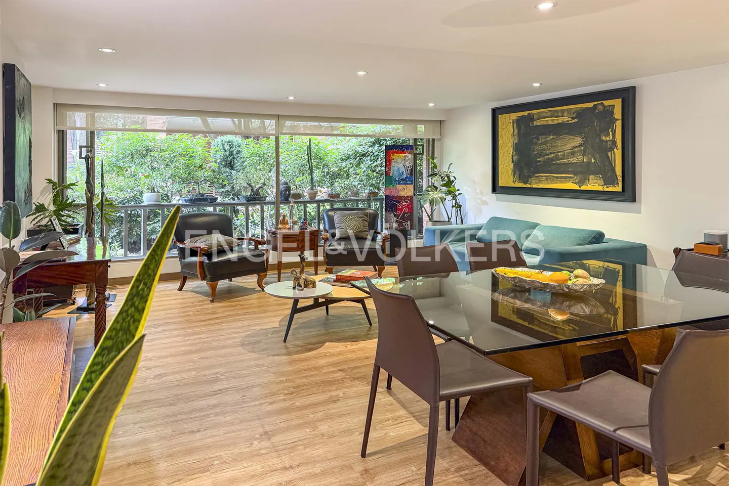 Bright living room with wood floors, a glass table, brown chairs, and a blue sofa. Large windows overlook lush greenery. Artwork adorns the walls.