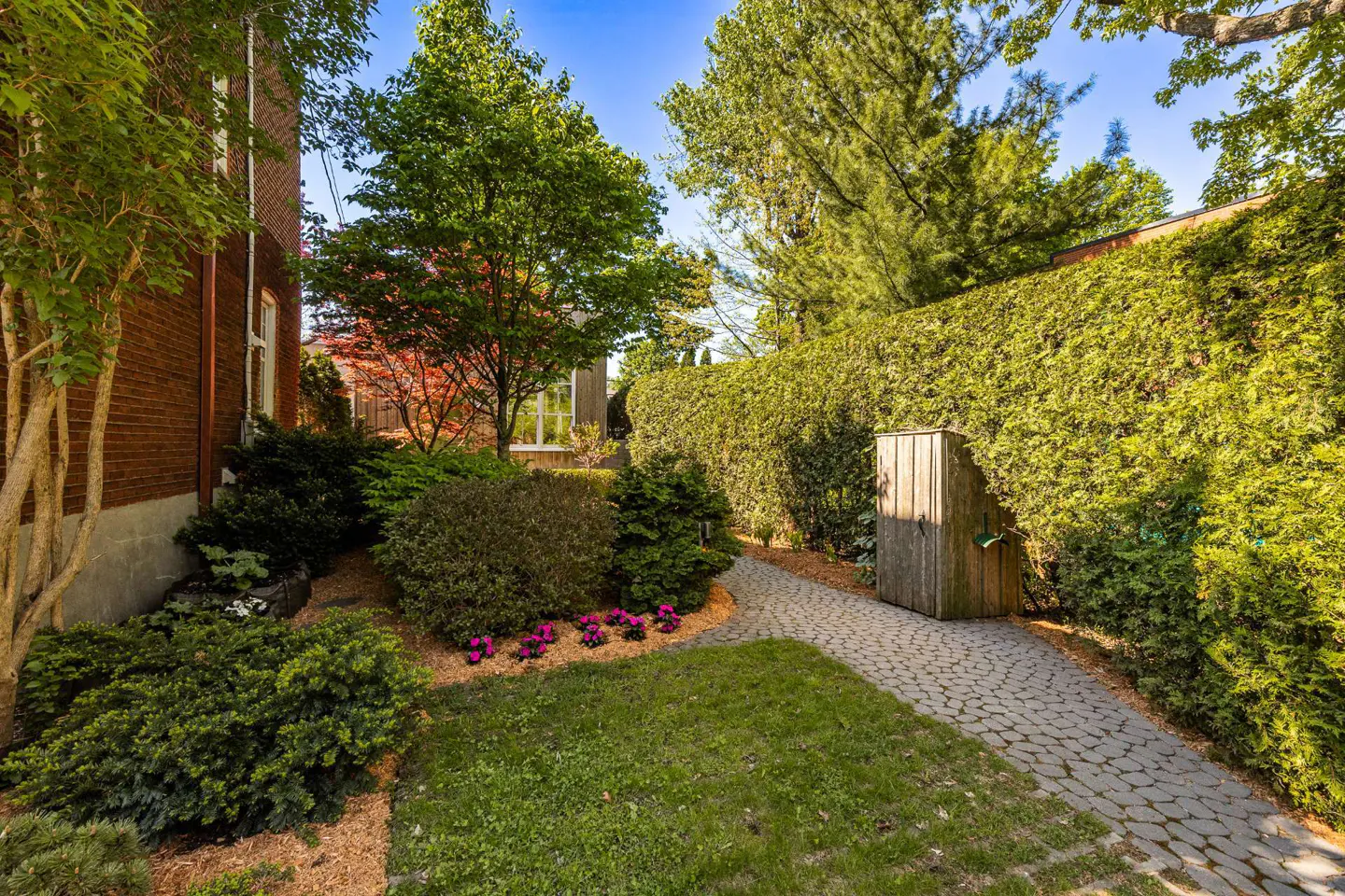 A brick house with a stone path leading to a tall green hedge and a wooden structure. The yard has grass, bushes, and flowers.