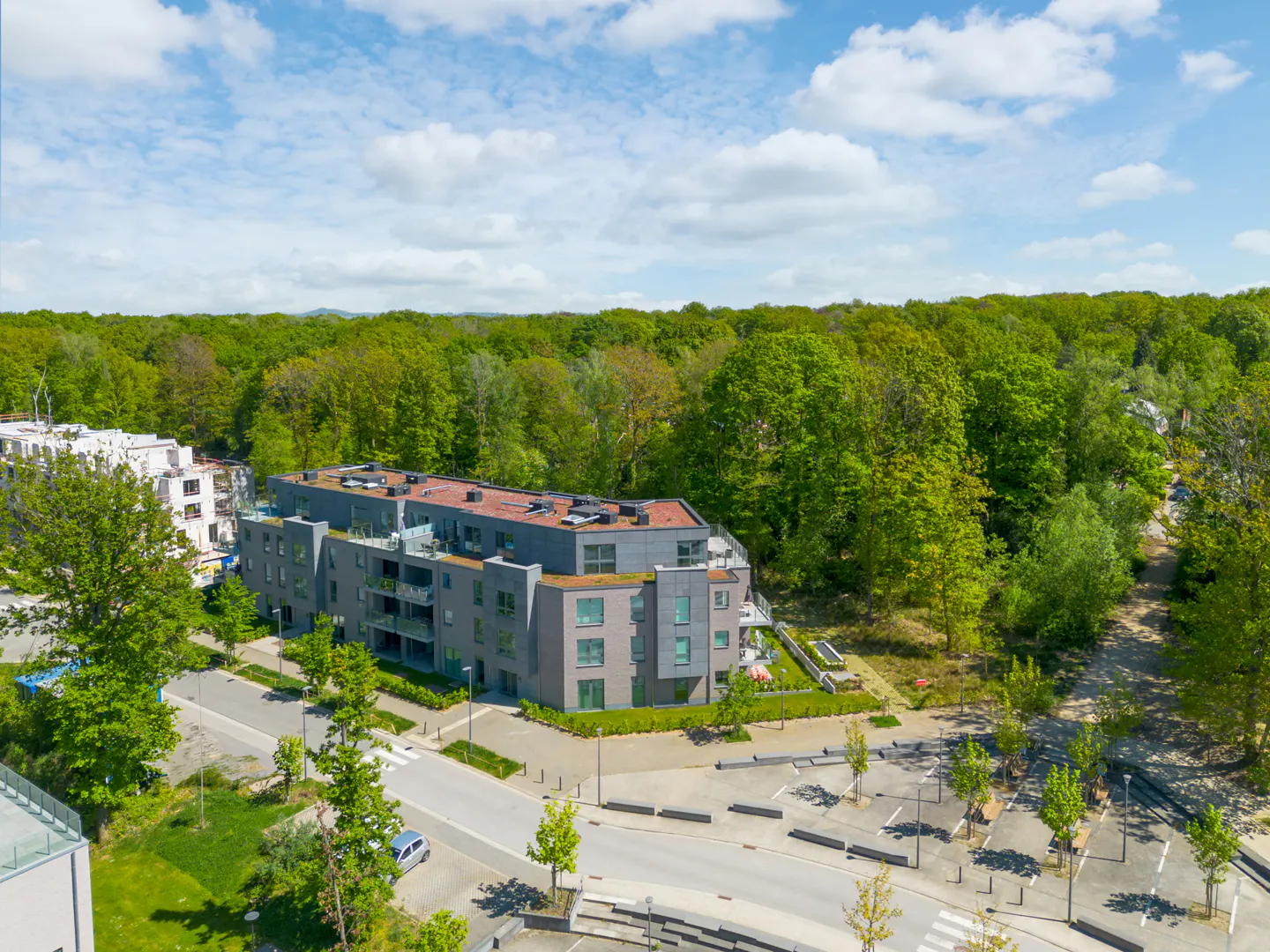Aerial view of a modern gray apartment building surrounded by green trees and a parking lot under a blue sky.
