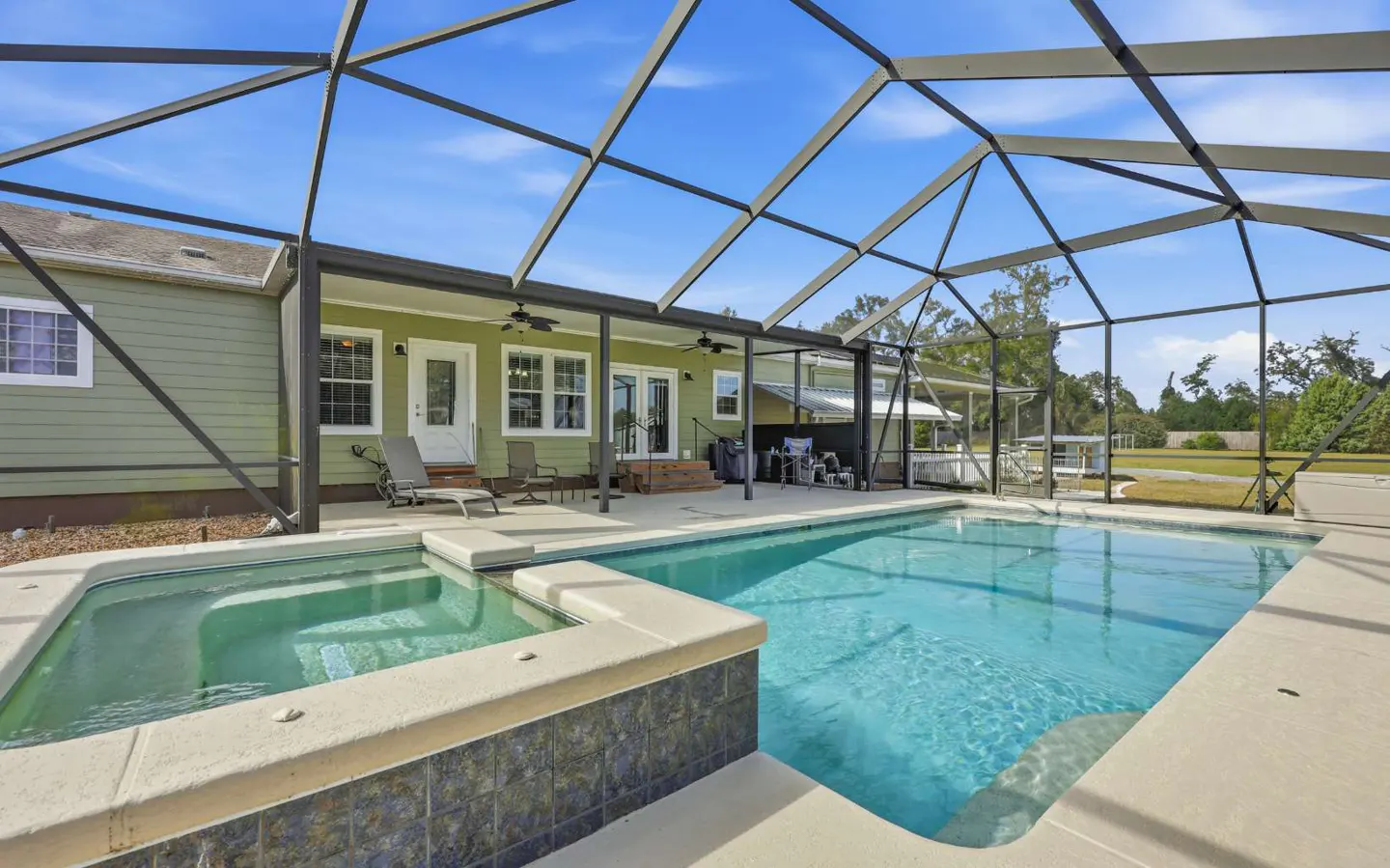 A backyard pool and hot tub are enclosed by a screen. The house is green with white trim. Blue sky is visible.