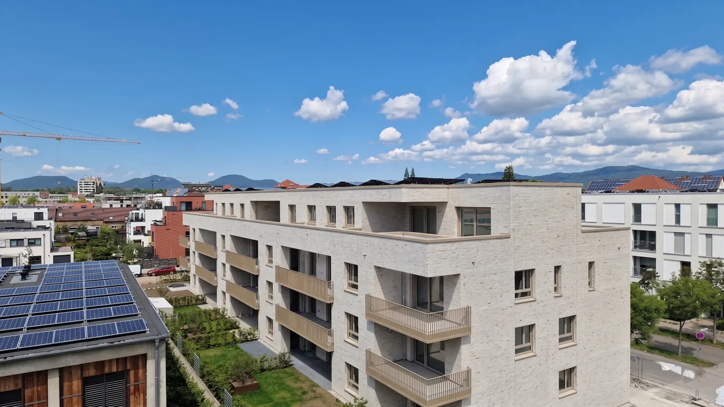 Modern apartment building with balconies, set against a blue sky with fluffy white clouds and distant mountains.