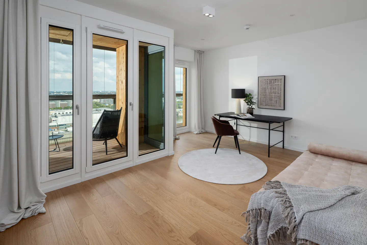 Bright room with wood floors, white walls, and a desk. Balcony with a chair is visible through the glass doors. A daybed is in the foreground.