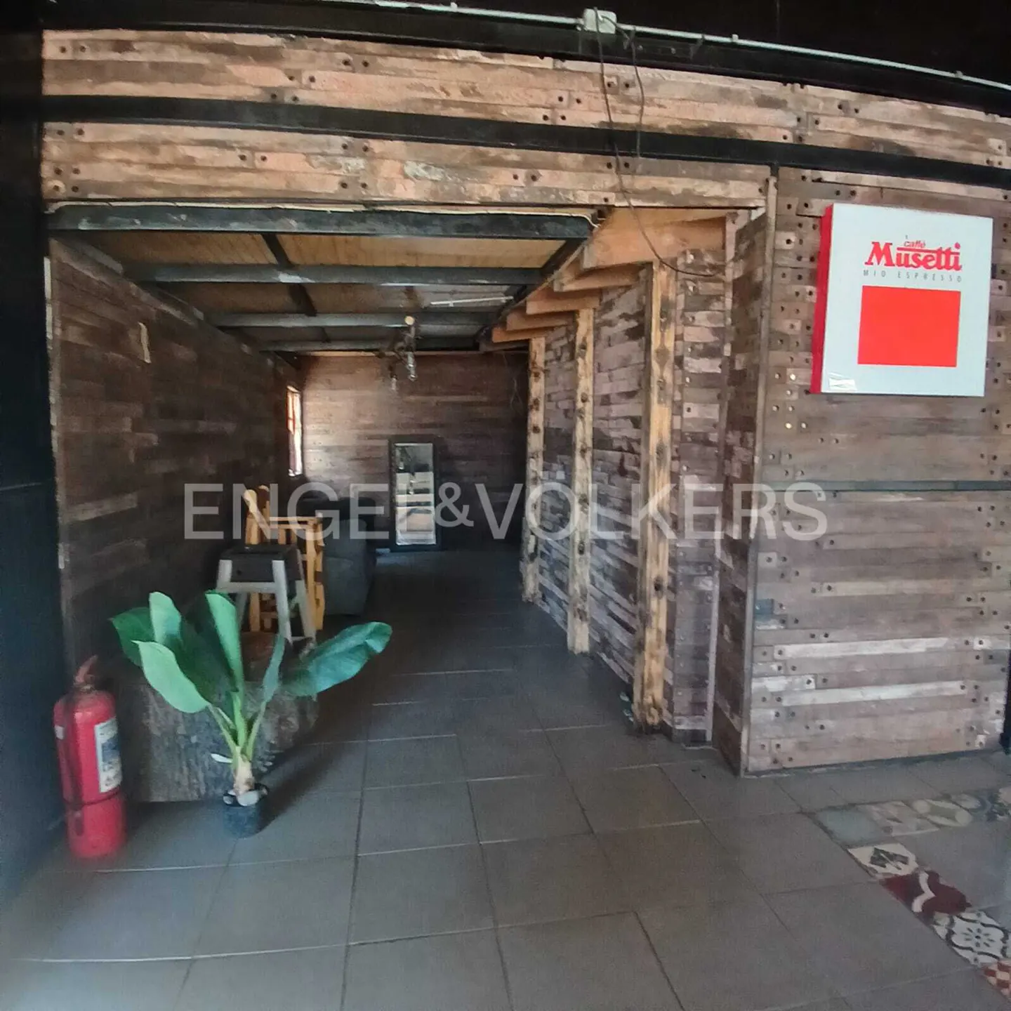Interior view of a rustic space with wood walls, tiled floor, and a "Musetti" sign. A plant and fire extinguisher are in the foreground.