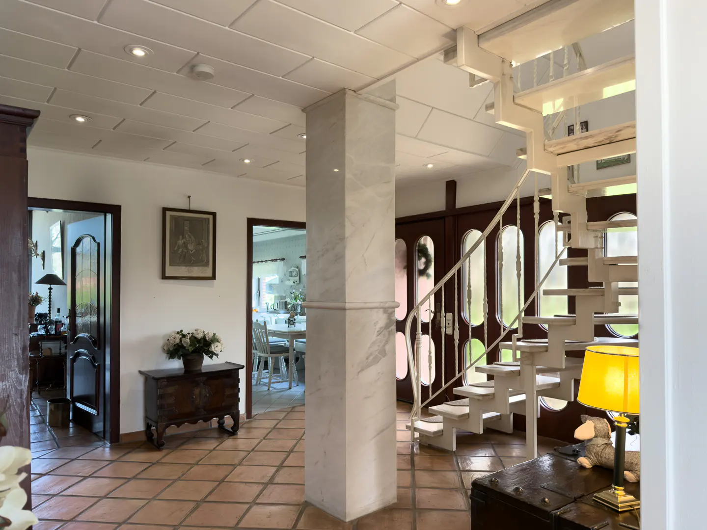 Interior view of a home with terracotta tile floors, white walls, and a white spiral staircase. A marble column stands near the stairs.
