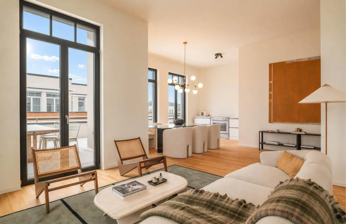 Bright, modern living room with hardwood floors, white walls, and black-framed windows. A white sofa, woven chairs, and a dining area with a glass table are visible.