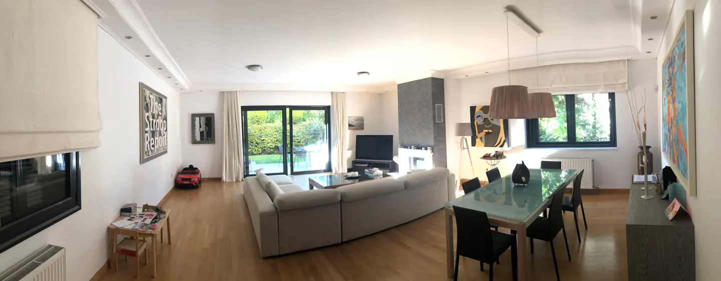 A bright, open-concept living and dining area with light wood floors and white walls. A large sectional sofa faces a TV and fireplace. A glass-topped dining table sits under pendant lights.