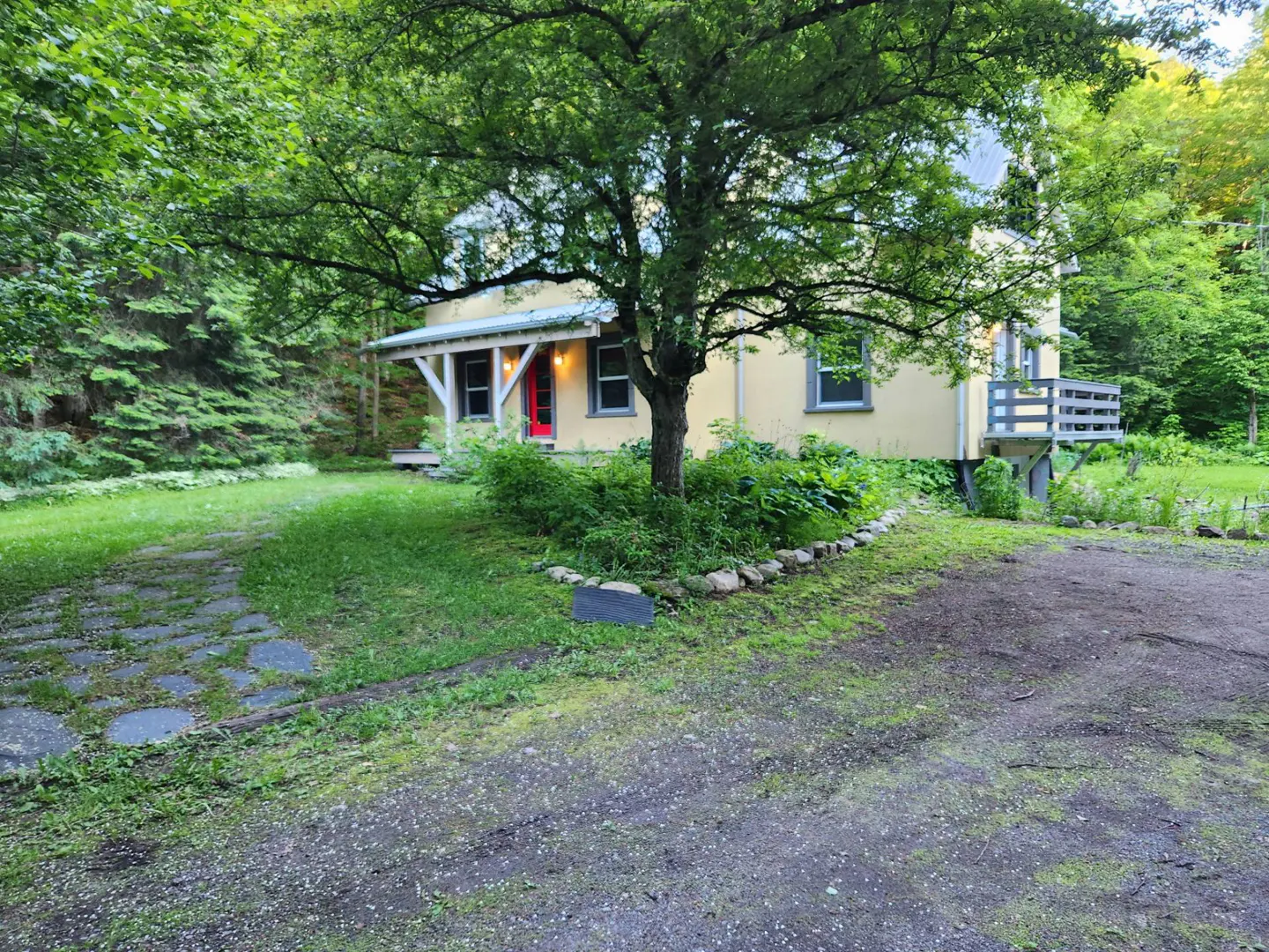 A yellow two-story house with a red front door, surrounded by lush trees and a stone path in a wooded setting.