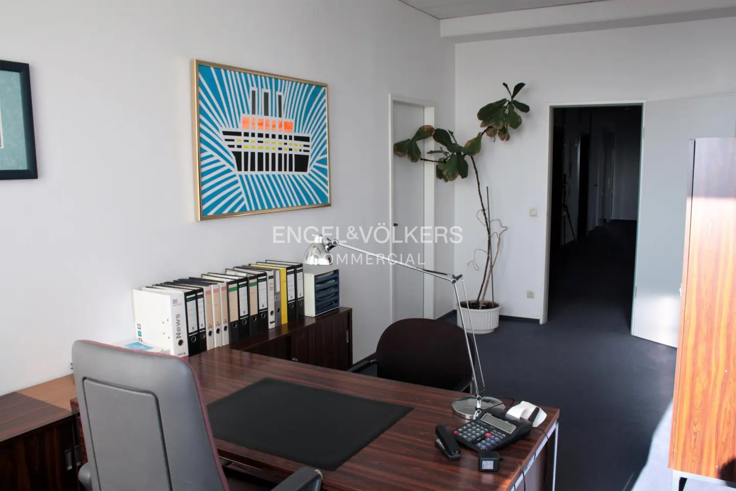 Office interior with a dark wood desk, gray chair, desk lamp, and a painting of a ship on the white wall. A plant sits near an open doorway.