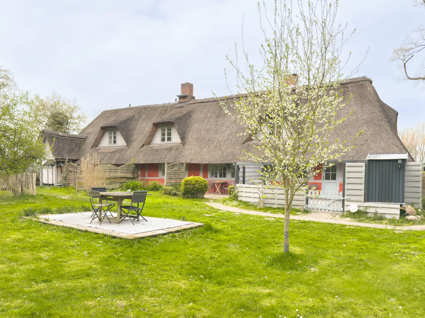 Exterior view of a thatched-roof house with a green lawn, a patio set, and a flowering tree in the yard.