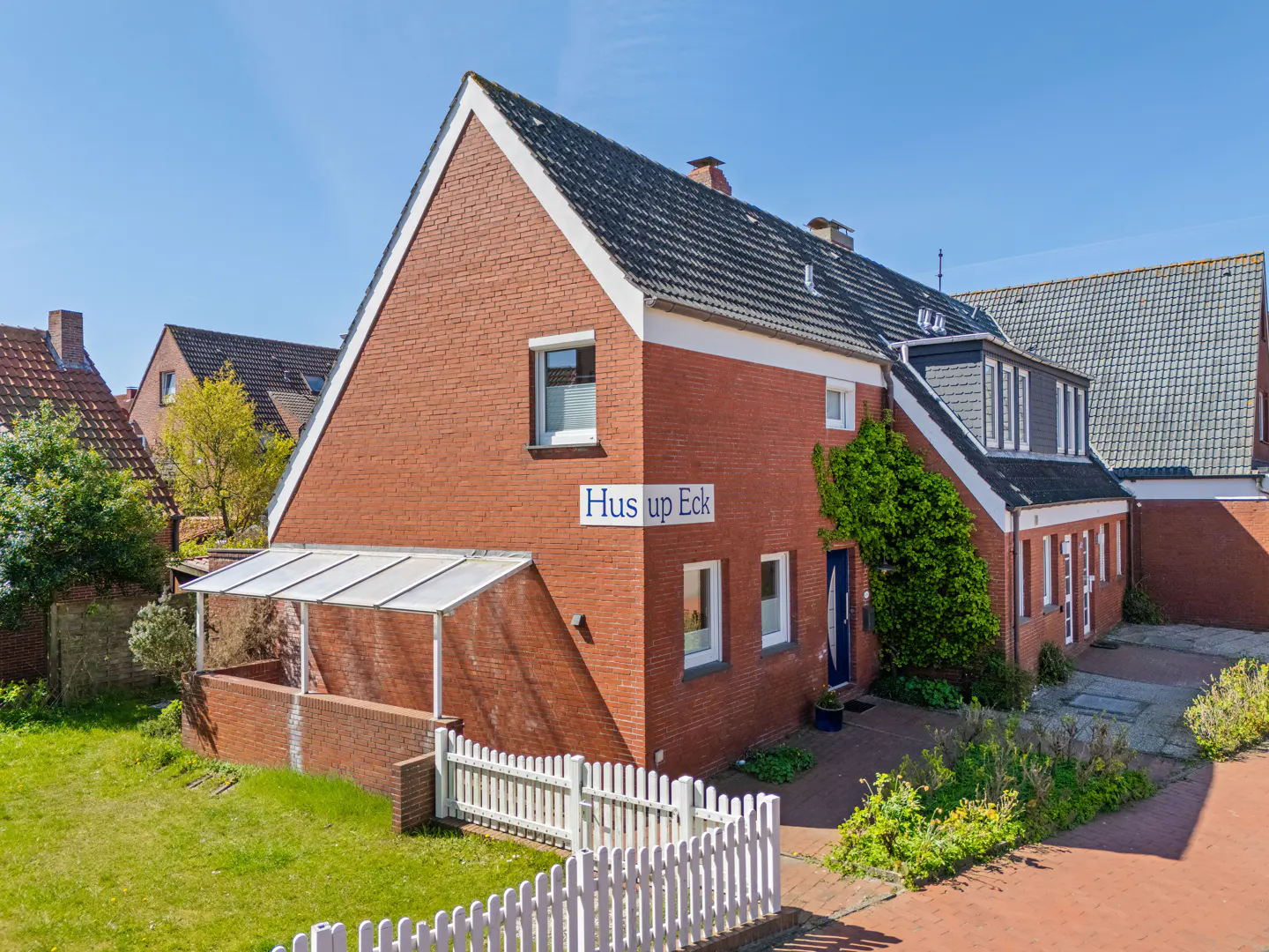 Exterior view of a two-story red brick house with a gray roof and white trim, named "Hus up Eck." A white picket fence borders the green lawn.