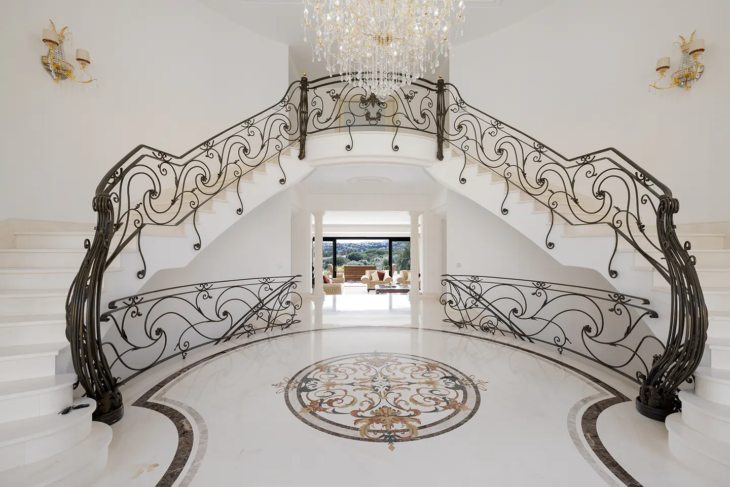 Elegant foyer with white marble floors, a decorative medallion, and a curved staircase with ornate wrought iron railings. A crystal chandelier hangs above.