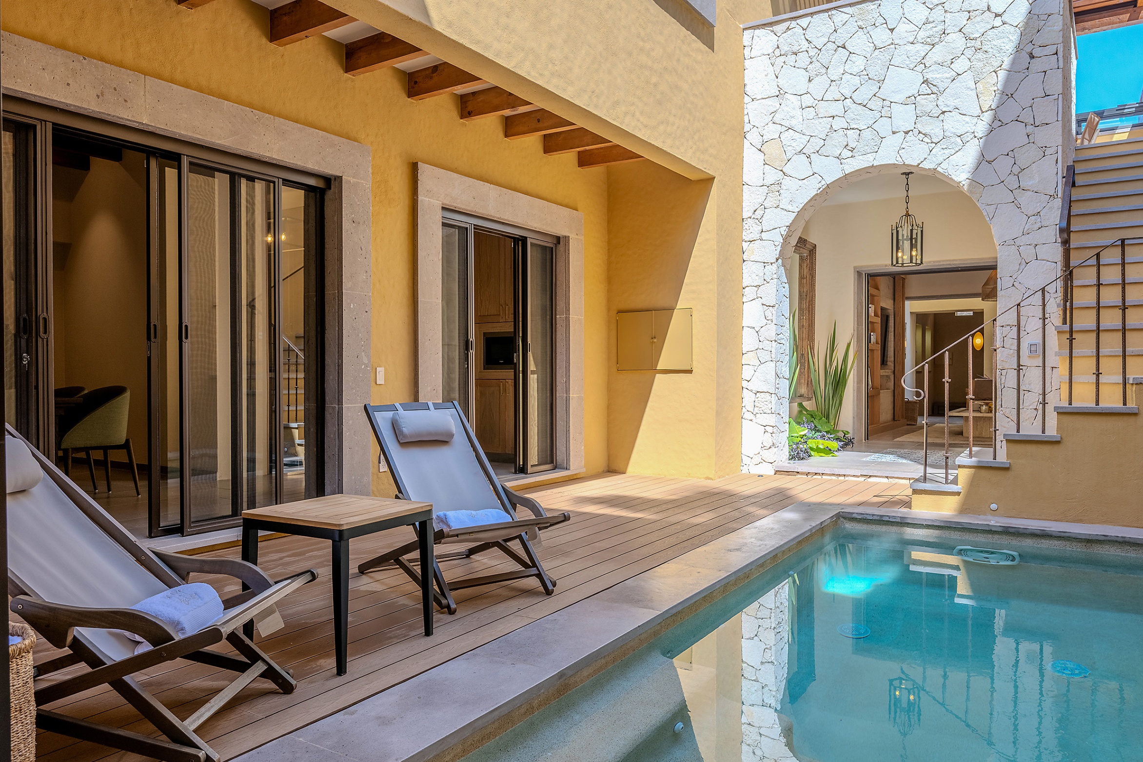 A sunny patio with a pool, wooden deck, two lounge chairs, and a stone archway leading to an interior living space.