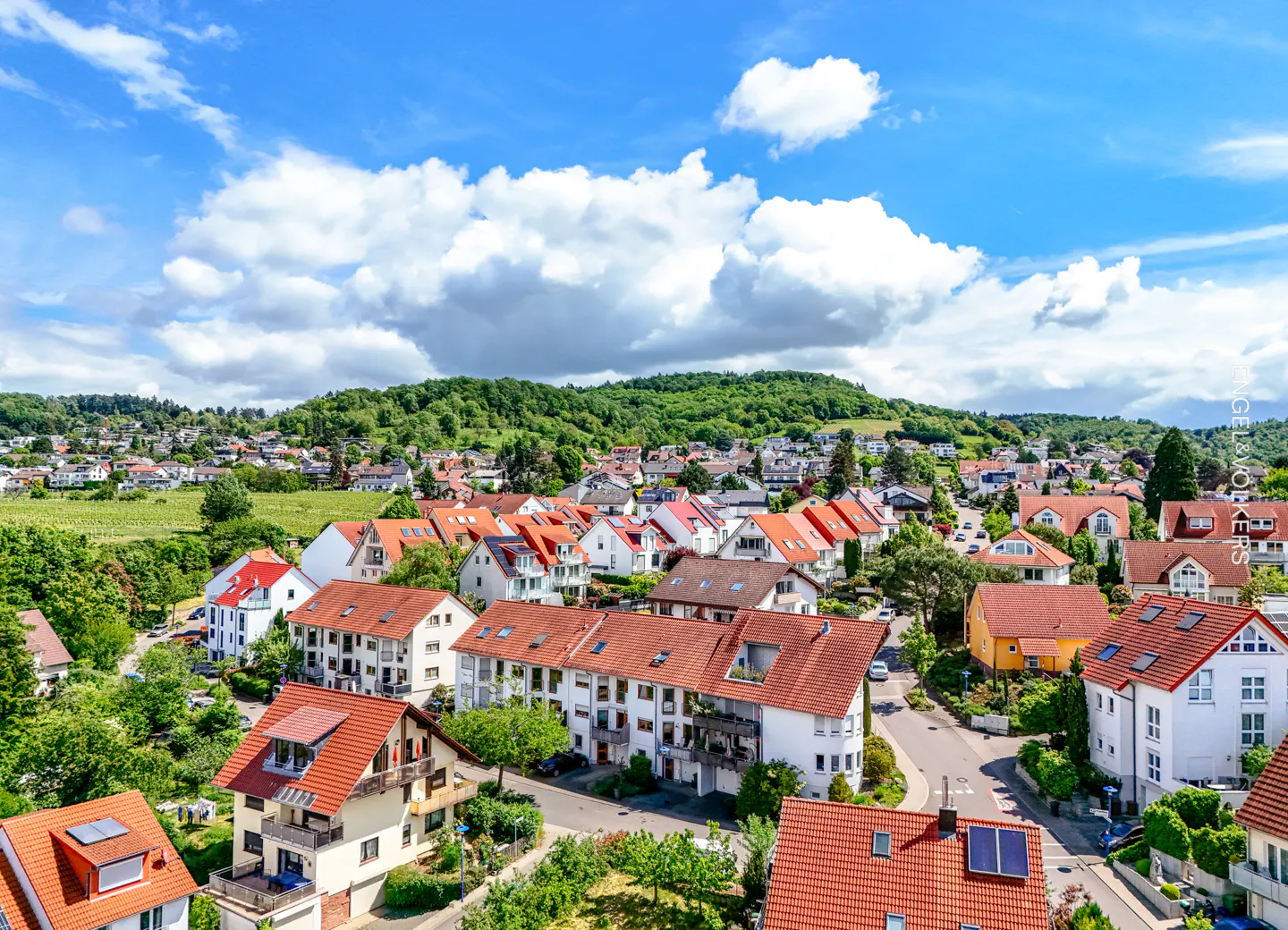 Aerial view of a residential area with red-roofed houses, green trees, and a blue sky with white clouds.