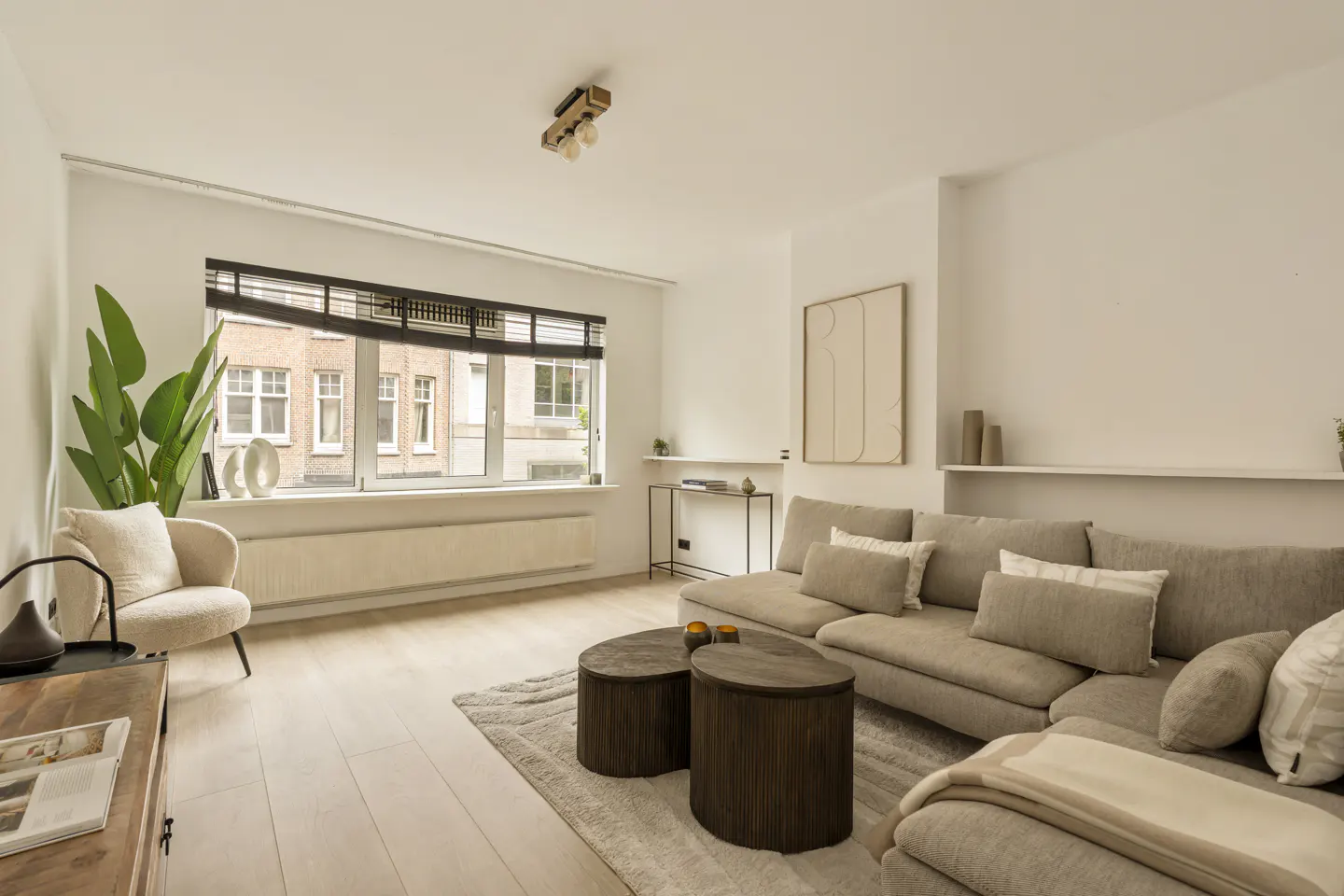 Bright living room with a beige sectional sofa, two dark wood coffee tables, and a cream armchair near a window.