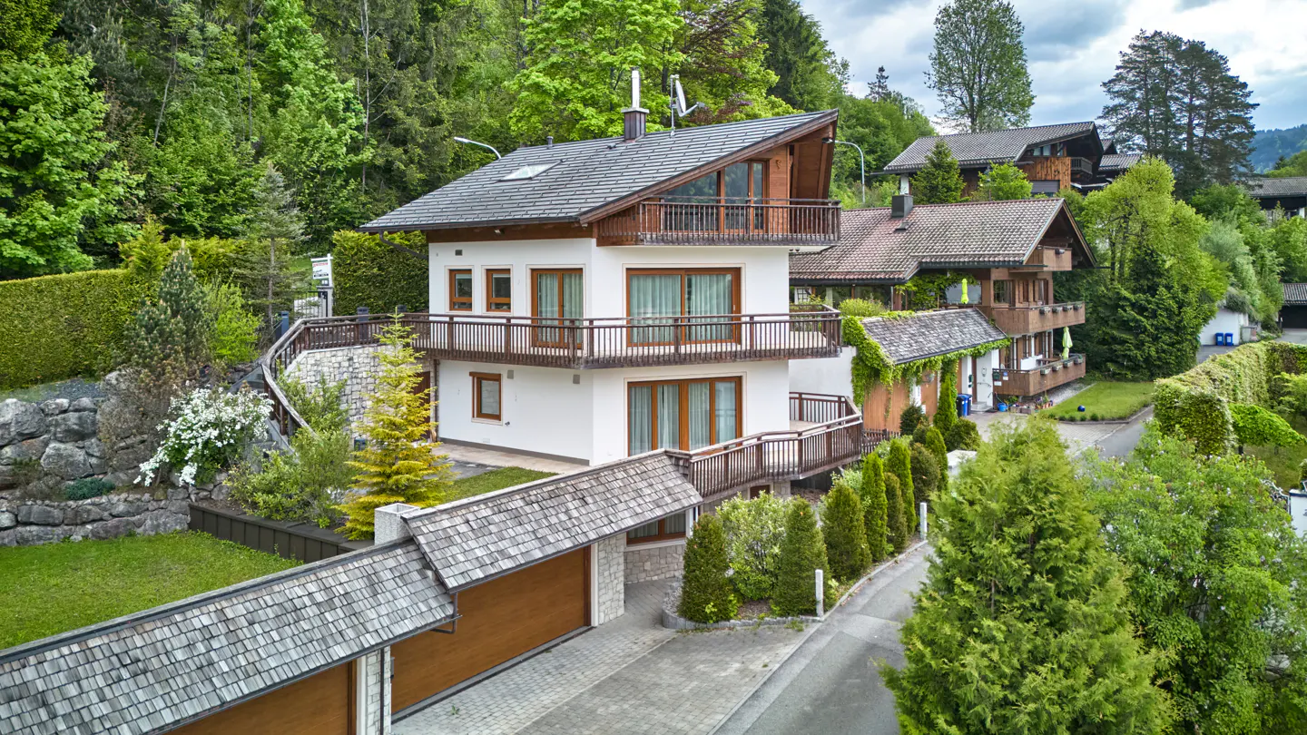 A modern white house with brown trim and balconies, nestled among green trees on a hillside.