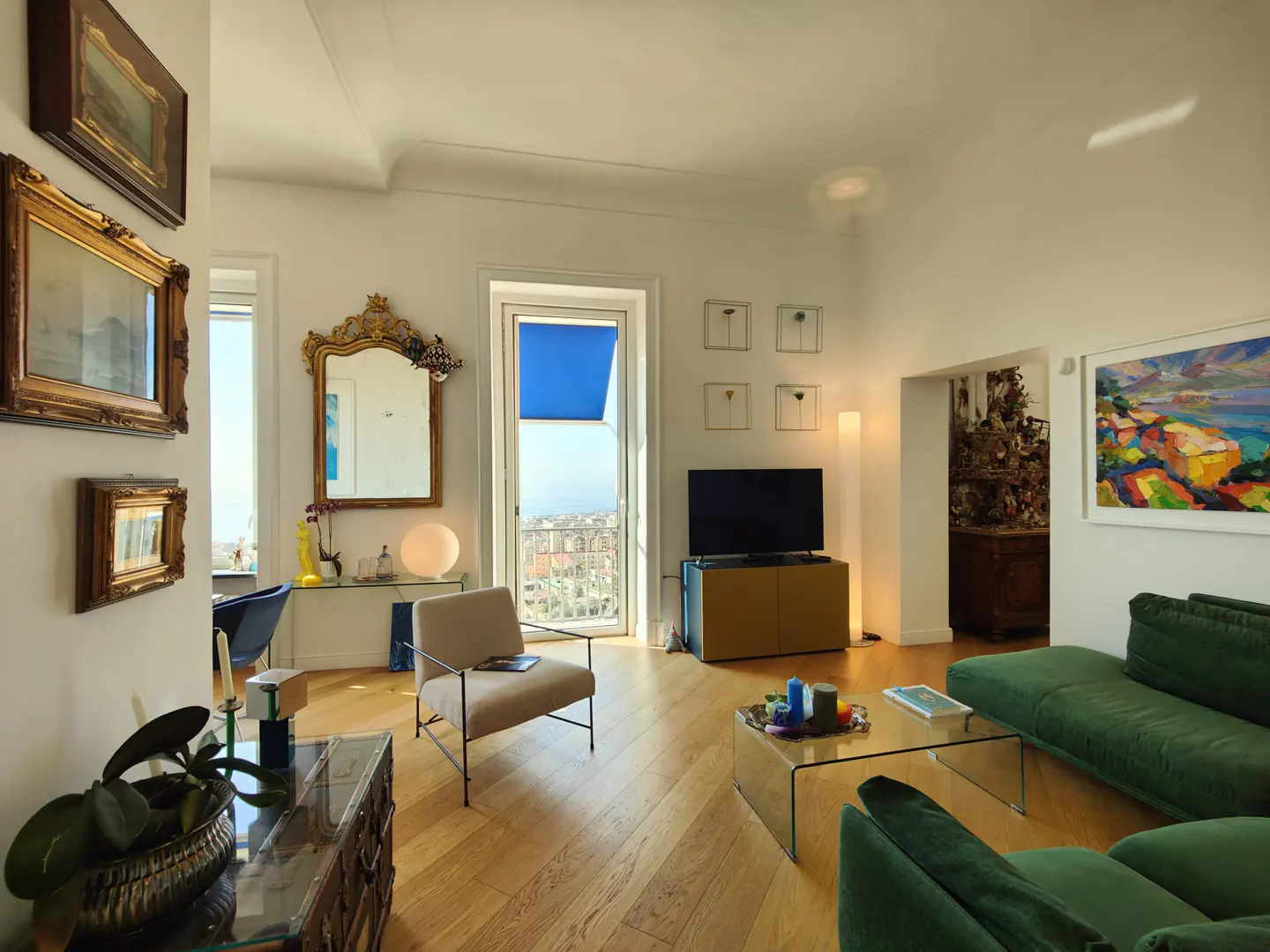 Bright living room with hardwood floors, green sofa, glass coffee table, TV, and balcony with a blue awning. Artwork adorns the walls.