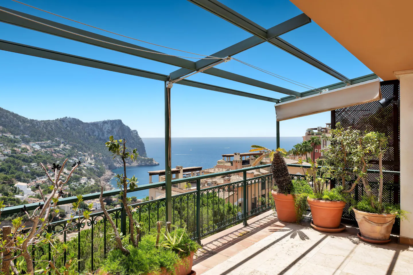 Terrace with potted plants and metal pergola overlooking a coastal town, rocky cliffs, and the blue Mediterranean Sea.