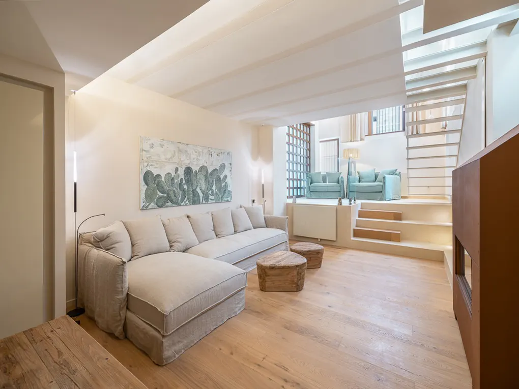 Bright living room with a beige sectional sofa, wooden coffee tables, and a cactus painting. Stairs lead to a seating area with two blue armchairs.
