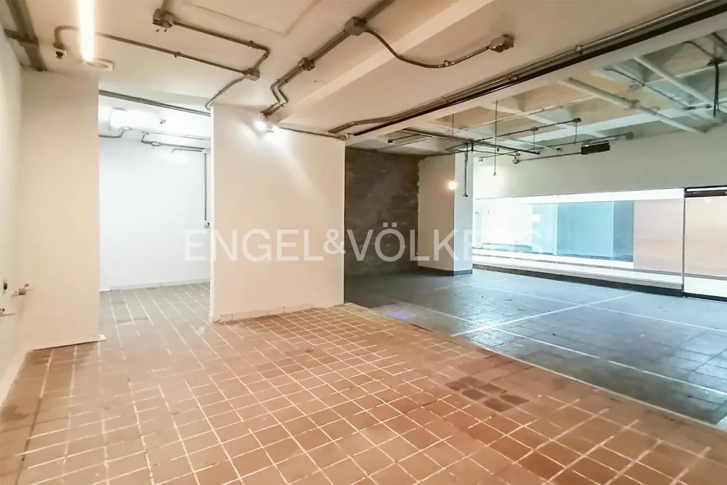 Spacious room with terracotta tile floor, white walls, and exposed ceiling pipes. Engel & Völkers logo visible on a wall.