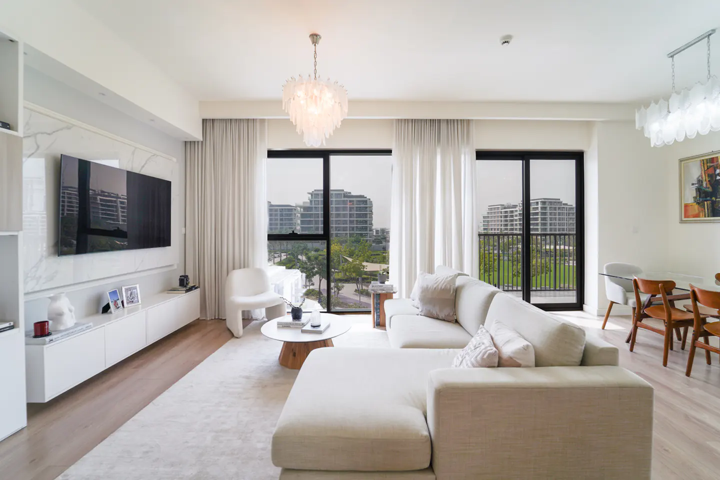 Bright living room with white sectional sofa, wood floors, and large windows overlooking city buildings and greenery. Dining table with chairs visible.