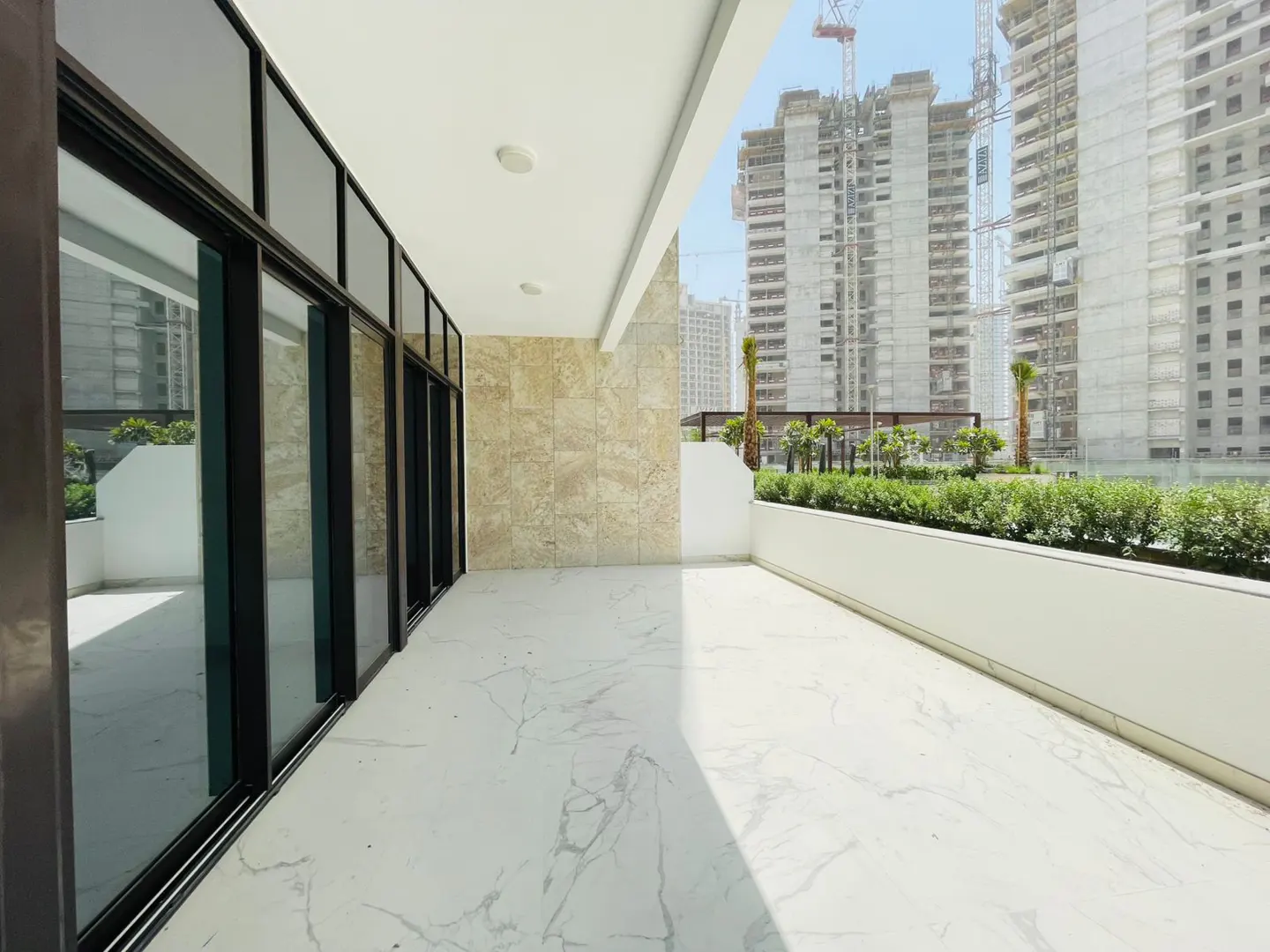 A bright, modern balcony with marble floors, black-framed glass doors, and a view of buildings under construction.
