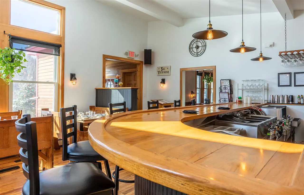 Interior view of a restaurant with a curved wooden bar, black stools, and pendant lights. A large window lets in natural light.