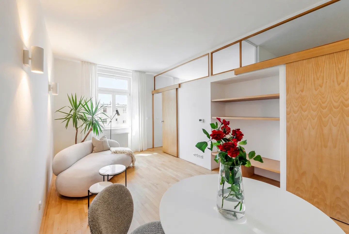 Bright, modern living room with hardwood floors, white walls, and a curved white sofa. Red roses in a vase sit on a white table. A large window lets in natural light.